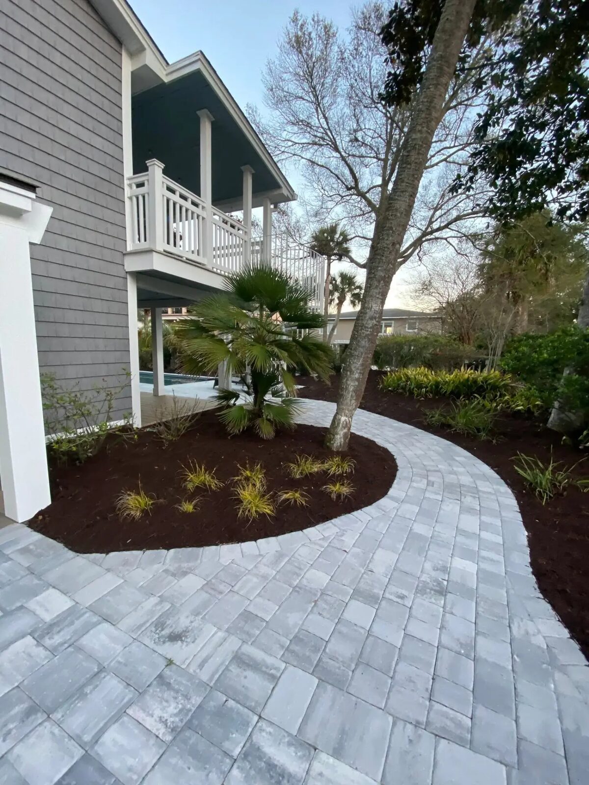 Curved grey paver walkway wrapping a coastal home with fresh dark mulch beds